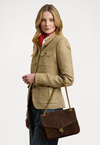 Geantă din suede maro cu un bretele din lanț auriu, având închiderea cu clapetă și logo-ul „RL” embosat, asortată cu un sacou bej cu model herringbone.