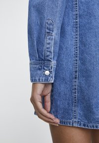 Robe en denim avec un effet délavé bleu, dotée d'un poignet à boutons et de détails sur la couture latérale. Texture lisse et coupe ajustée.