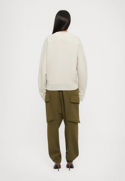 Woman with long dark hair wearing a beige oversized sweatshirt and olive green cargo pants with large pockets, facing away on a white background.