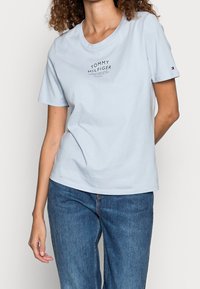 Camiseta de algodón de color azul claro con cuello redondo y mangas cortas. En la parte frontal presenta el logo "TOMMY HILFIGER" en texto negro.