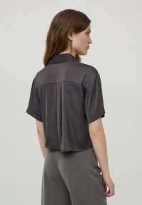 Camicia grigia a maniche corte in tessuto liscio, caratterizzata da un colletto e una vestibilità ampia con una cucitura centrale sulla schiena. Indossata con pantaloni grigi.