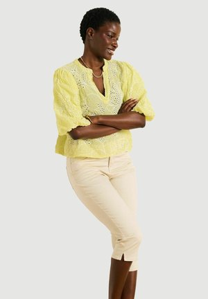 Kobieta w żółtej bluzce z ażurowym wzorem i beżowych krótkich spodniach, stojąca z założonymi rękami i uśmiechająca się na jednolitym tle.