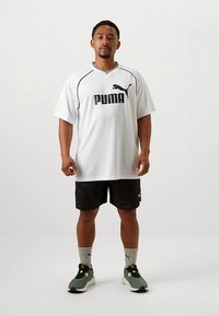 Mees, kes seisab valges taustal, seljas valge Puma T-särk, mustad lühikesed püksid, hallid Puma sokid ja rohelised spordijalatsid.