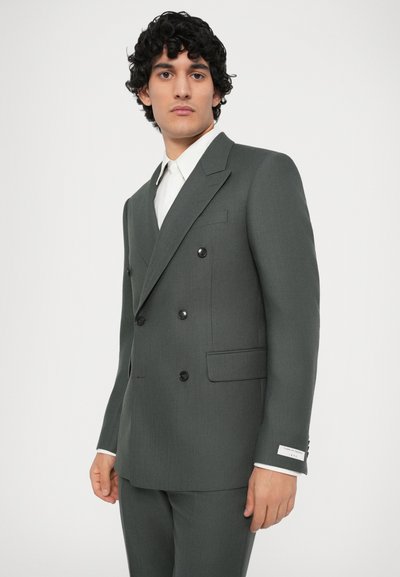 Abito grigio doppiopetto con tessuto testurizzato. Presenta bottoni neri e una vestibilità su misura. Camicia bianca visibile al colletto e ai polsini.