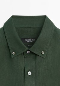 Mörkgrön linneskjorta med button-down-krage och vita knappar, med en etikett med varumärke och tygdetaljer inne i kragen.