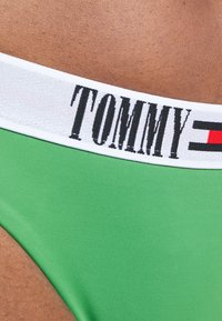 Zelené plavky s hladkým povrchem, vybavené bílým elastickým pasem s nápisem "TOMMY" v tučném černém písmu a červenými akcenty.