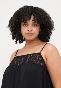 Haut sans manches noir avec des détails en dentelle, doté de bretelles fines et d'un tissu fluide. La dentelle présente un motif géométrique le long de l'encolure.