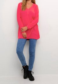 Blusa rosa a maniche lunghe con scollatura a V, abbinata a jeans skinny blu e stivali alla caviglia neri con lacci. Tessuto liscio, vestibilità casual.