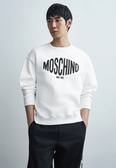 Λευκό φούτερ με μαύρο λογότυπο «Moschino», με ριμπ μανσέτες και τελείωμα, συνδυασμένο με σκούρο παντελόνι, στέκεται μπροστά σε ανοιχτό φόντο.