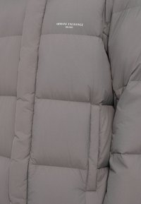 Sivá prešívaná bunda s hladkou textúrou a prednou vreckom. Výrazne zobrazené logo "Armani Exchange Milano".