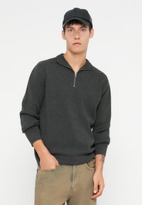 Maglione zip grigio scuro realizzato in tessuto testurizzato con una vestibilità rilassata, abbinato a jeans beige chiaro e un cappellino nero.