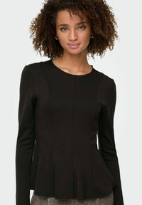 Kvinde med krøllet hår iført en tætsiddende sort langærmet bluse med peplum kant, stående mod en ensfarvet lys baggrund.