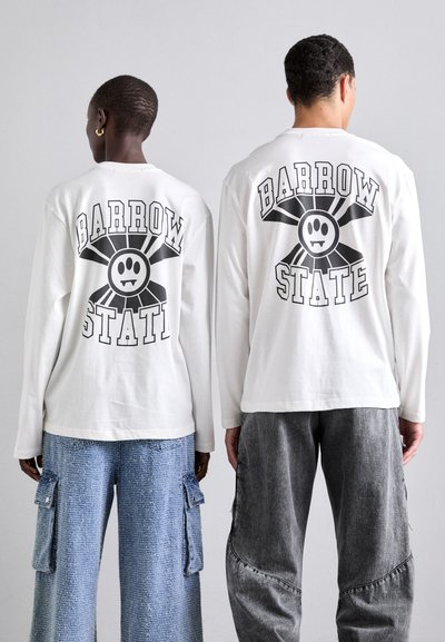 Chemises blanches à manches longues avec un graphique "BARROW STATE" et un design de visage souriant au dos; associées à un pantalon cargo bleu et gris.