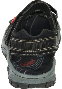 Svart idrottssko med övre del av texturerad tyg, grå gummisula, justerbar rem och röd logotypdetalj. Bakifrån visas skons form.