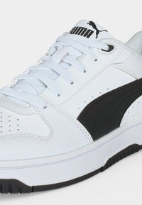 Λευκό αθλητικό παπούτσι Puma με μαύρη λωρίδα λογότυπου, λευκά κορδόνια, διάτρητη μύτη και μαξιλαρωμένη σόλα σε απλό φόντο.