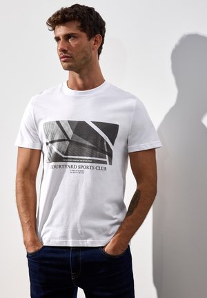 Hvid bomulds T-shirt med et geometrisk sort og gråt grafisk tryk, med teksten "COURTYARD SPORTS CLUB" i fede bogstaver nederst.