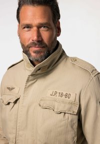 Giacca stile militare beige con collo alto, due tasche frontali e bottoni sulle spalle. Presenta dettagli con testo ricamato e un emblema.