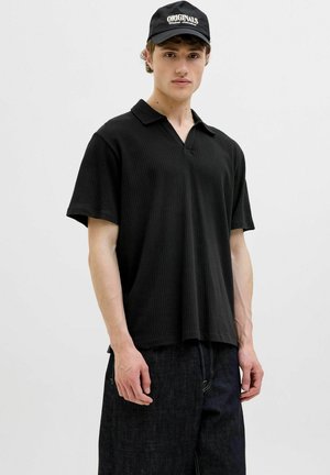 Black ribbed polo shirt with a relaxed fit and a short collar, paired with dark denim pants. A black cap with "ORIGINALS" logo is worn.