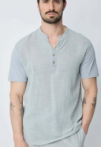 Chemise gris clair à manches courtes avec une finition texturée, dotée d'un col henley, de trois boutons en bois, et d'une coupe décontractée.