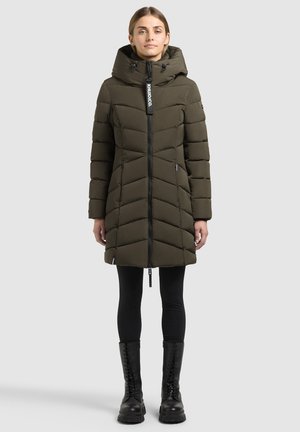 Femme portant un long manteau matelassé vert olive avec capuche, des leggings noirs et des bottes noires à lacets, debout devant un fond uni.