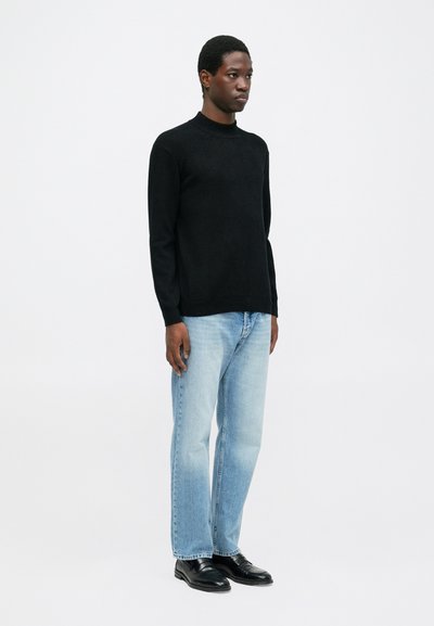 Uomo in piedi che indossa un maglione nero, jeans azzurri e scarpe eleganti nere su uno sfondo bianco uniforme.