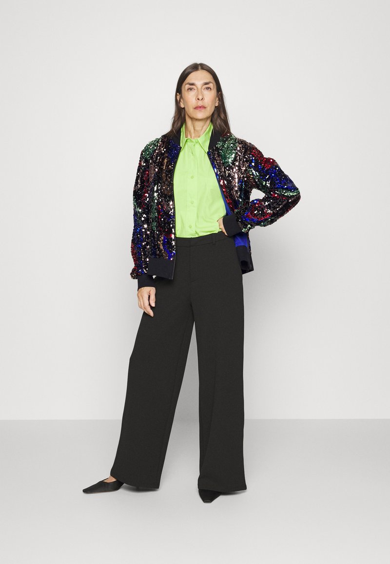 Veste bomber en sequins avec des paillettes multicolores, associée à une chemise vert vif à boutons et un pantalon large noir. Chaussures plates noires.