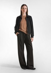 Donna in piedi con le mani in tasca, che indossa una giacca nera con zip, una camicia marrone, pantaloni a vita alta a righe sottili scuri e scarpe nere.