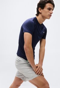 Νέος άνδρας τεντώνοντας τα πόδια του, φορώντας μπλε αθλητικό μπλουζάκι και ανοιχτό γκρι σορτς, μπροστά σε απλό λευκό φόντο.