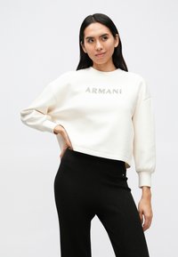 Λευκό κοντό φούτερ με διακριτικό στρογγυλό λαιμό και μακριά φουσκωτά μανίκια· διαθέτει λογότυπο «ARMANI» σε ασημί χρώμα στο στήθος. Μαύροι ριμπ κολάν.