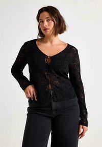 Cardigan in pizzo nero con maniche lunghe, scollo a V profondo e chiusura con bottoni con dettaglio a occhiello. Indossato sopra jeans scuri.