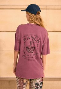 Unisex överdimensionerad mauve T-shirt med en svart ramen-grafik och text på baksidan; kombinerad med mönstrade byxor i jordnära toner.