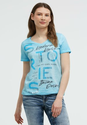 Junge Frau mit langen Haaren, die ein blaues T-Shirt mit Grafik und Text sowie blaue Jeans trägt, steht vor einem einfarbigen hellen Hintergrund.