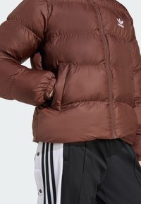 Brun pufferjakke med en glat, skinnende tekstur og lynlås lukning. Har elastiske manchetter og et Adidas-logo på brystet. Stylet med sorte bukser.