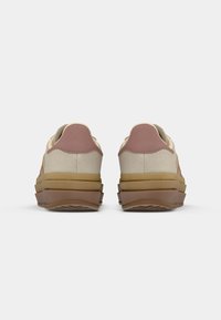 Des baskets en tissu beige présentent une semelle châtaigne avec une texture antidérapante, des accents roses au talon et un rembourrage superposé pour plus de hauteur.