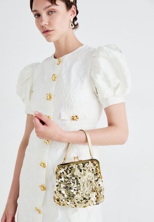 Mujer vistiendo un vestido blanco con mangas abullonadas texturizadas y botones de flor dorados, sosteniendo un pequeño bolso de mano dorado con lentejuelas y asa superior.