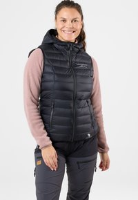 Svart puffy väst med huva, dragkedjestängning och logotyp på bröstet. Har en quiltad design och är lager-på-lager över en rosa långärmad topp.