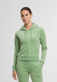 Zöld velúr kapucnis pulóver, elöl zipzárral, kapucnival és elülső zsebekkel. Arany színű fém kiegészítőkkel és egy kis hímzett logóval a mellkasán.