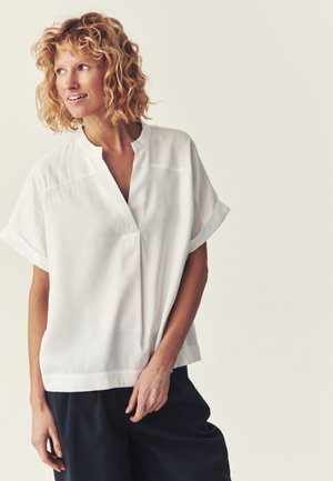 Vrouw met krullend blond haar, een losse witte blouse met korte mouwen en een donkere broek, kijkt naar links tegen een effen lichte achtergrond.