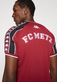 Κόκκινη αθλητική φανέλα με το γράψιμο FC METZ, μαύρες λεπτομέρειες και λογότυπο Kappa στους ώμους, με κοντά μανίκια και σχέδιο με στρογγυλή λαιμόκοψη.