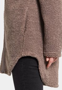 Ruskea fleece-takki, jossa on teksturoitu, pehmeä materiaali. Takissa on kaareva helma ja sivuvetoketju, ja se on kuvattu sivulta, käsi lepäämällä vaatteen päällä.