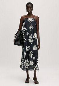 Schwarzes ärmelloses Kleid mit weißen, maritim inspirierten Mustern, fließendem Design und Midi-Länge. Begleitet von einer schwarzen Handtasche und Sandalen.