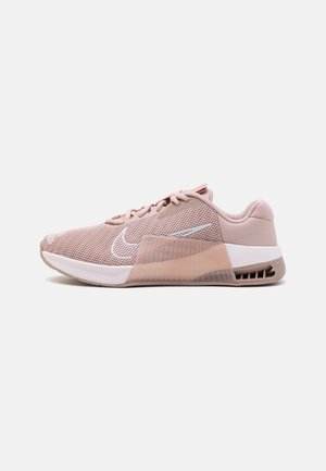 Zapatilla deportiva rosa claro con parte superior de malla, logo blanco de Nike, media suela blanca y soporte de plástico transparente en el lateral sobre un fondo liso.