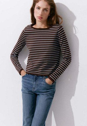 Jeune femme portant un t-shirt à manches longues rayé marron et blanc et un jean bleu, debout les mains dans les poches devant un mur blanc.