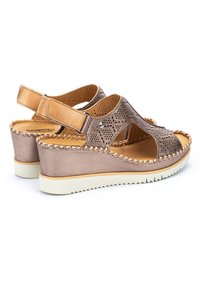 Sandalias cuña hechas de cuero bronce metálico perforado con una correa beige, que cuentan con una suela texturizada y costuras decorativas a lo largo de los bordes.