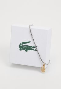 Brățară din lanț argintie cu un charm în formă de crocodil auriu, așezată pe o cutie texturată albă cu un logo crocodil verde.