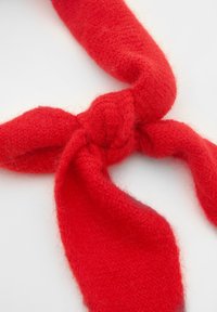 Červený vlněný šátek s měkkou, chlupatou texturou, uvázaný do uzlu s dvěma dlouhými, zužujícími se konci. Materiál vypadá silně a zachovává teplo.