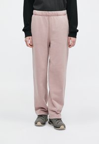 Άτομο που φοράει ανοιχτό ροζ βελούδινο παντελόνι με φαρδιά πόδια, μαύρη μακρυμάνικη μπλούζα και γκρι αθλητικά παπούτσια, να στέκεται μπροστά σε απλό φόντο.