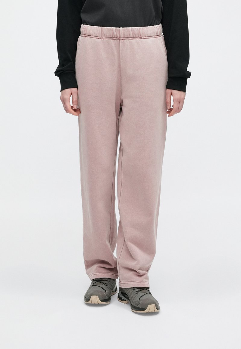 Persona che indossa pantaloni a zampa d'elefante di velluto rosa chiaro, una maglietta nera a maniche lunghe e sneakers grigie, in piedi contro uno sfondo neutro.