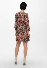 Žena stojící zády, oblečená v květinových šatech po kolena s dlouhými rukávy a světle zbarvenými kotníkovými botami na bílém pozadí.