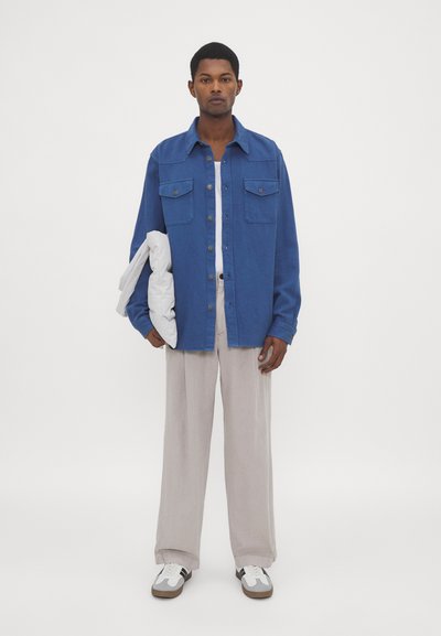 Giacca camicia di cotone blu con due tasche sul petto, abbinata a pantaloni ampî grigi chiari e sneakers bianche. Modella che tiene un oggetto bianco.